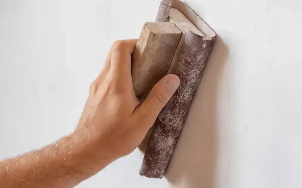 Mano lijando paredes para pintar. El bloque de madera con lija se desliza sobre la superficie blanca, preparándola para un acabado perfecto.